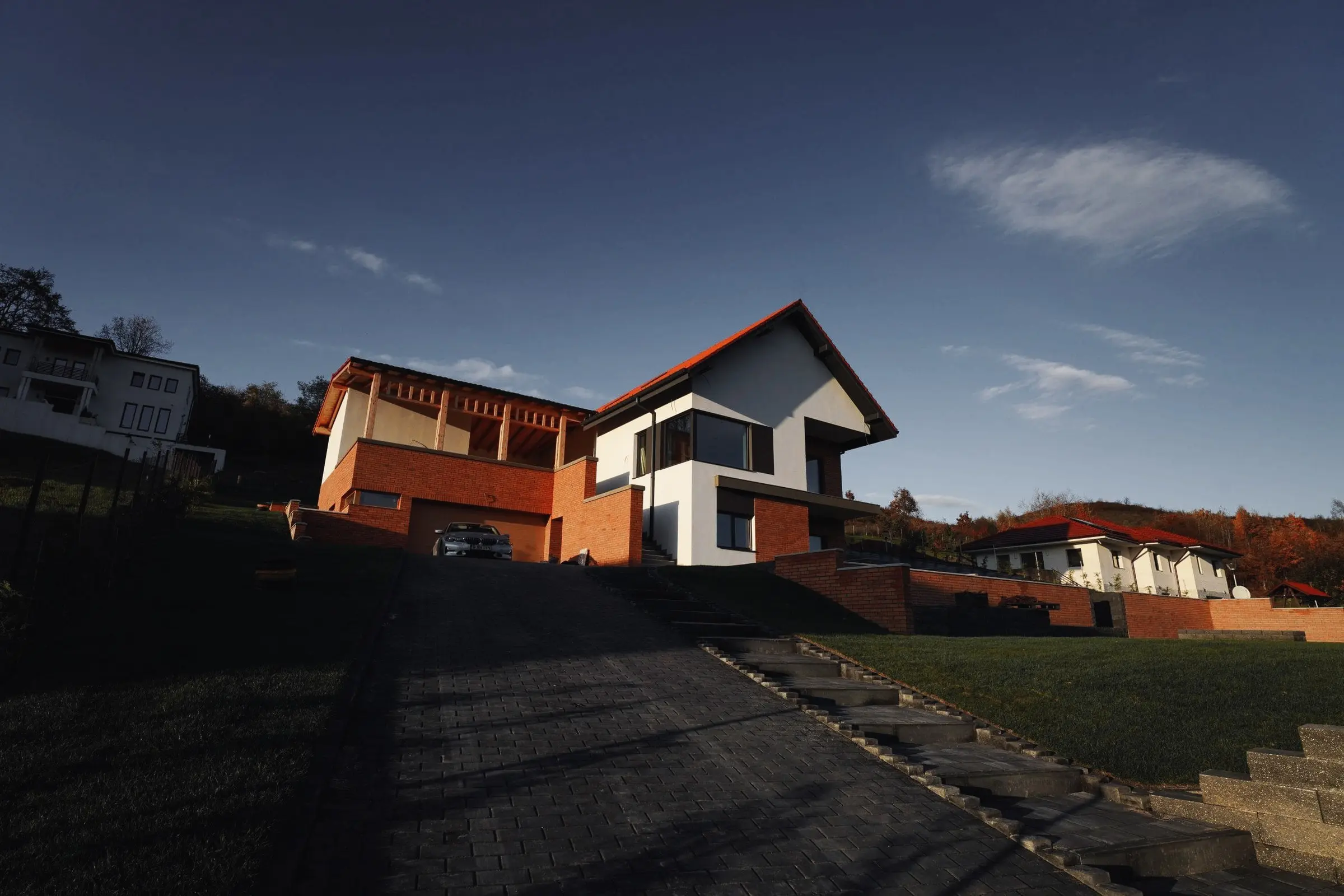 Casă modernă din cărămidă și tencuială albă, pe deal, cu alee pavată la apus
