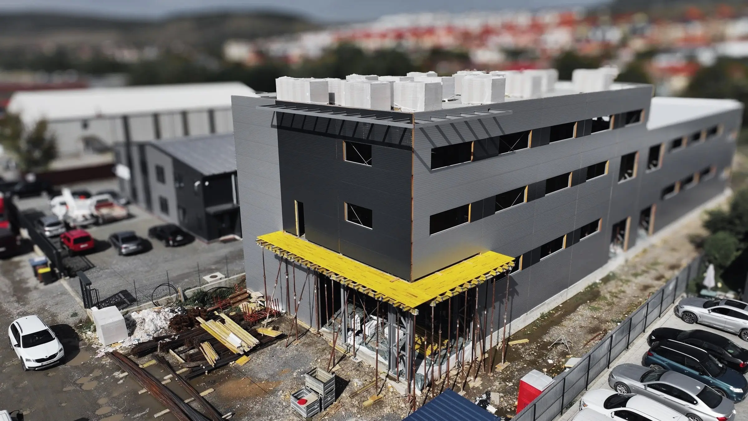 Clădire cu trei niveluri și fațadă gri în construcție, cu parcare alături, din dronă
