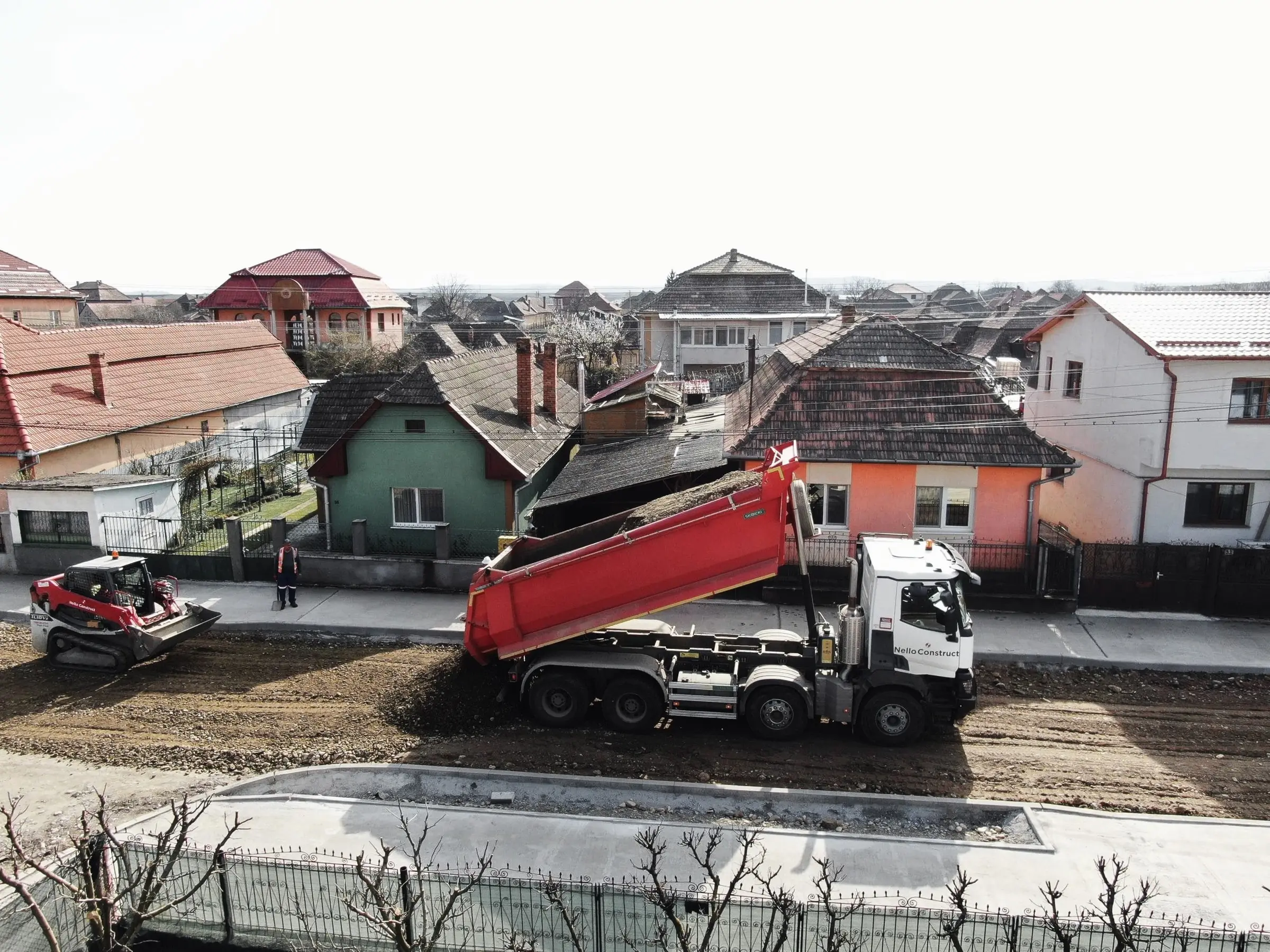 Autobasculantă descarcă material pe o stradă în lucru, între case, cu un compactor alături