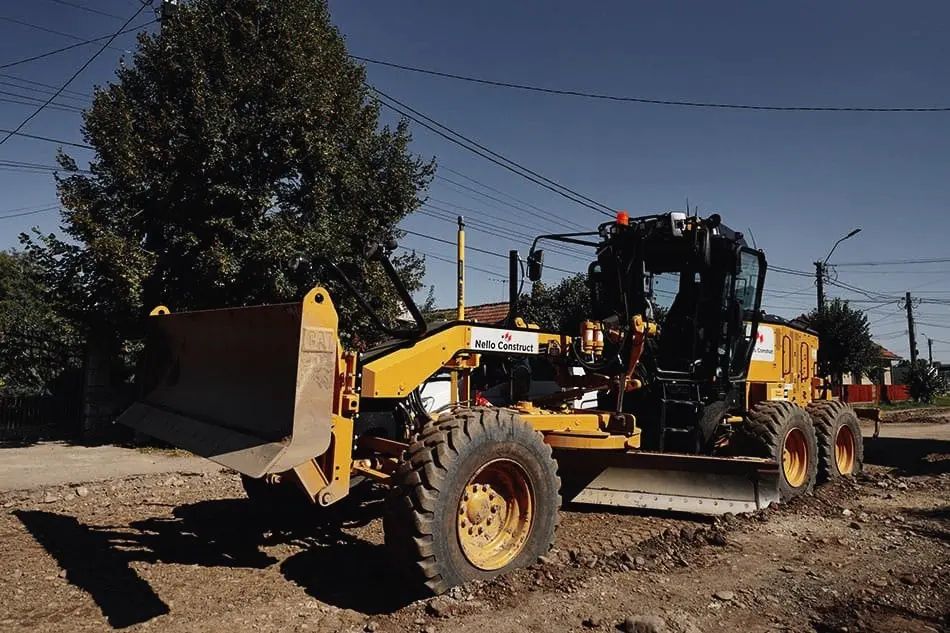 Autogreder galben nivelează o stradă de pământ, cu inscripția Nello Construct pe lamă