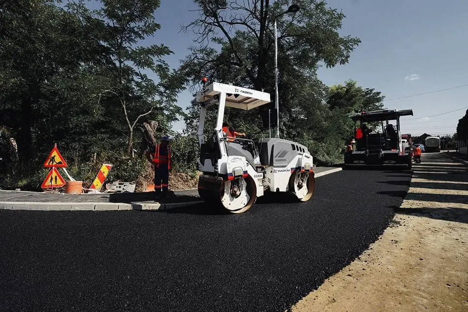 Compactor cu tambur și finisor lucrează la asfaltarea unui drum, cu muncitori pe margine