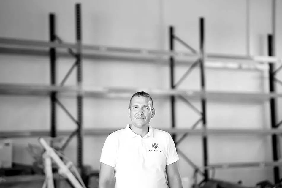 Bărbat în tricou alb cu sigla Nello Construct, în fața unor rafturi metalice roșii
