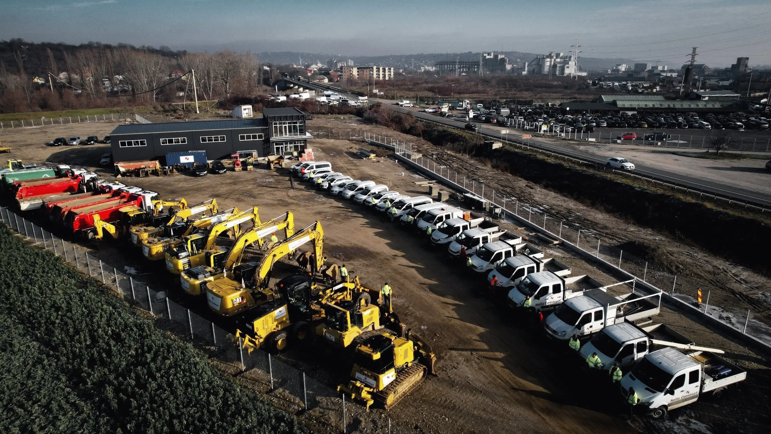 Utilaje galbene, autobasculante și autoutilitare parcate în rânduri pe un teren, văzute din dronă