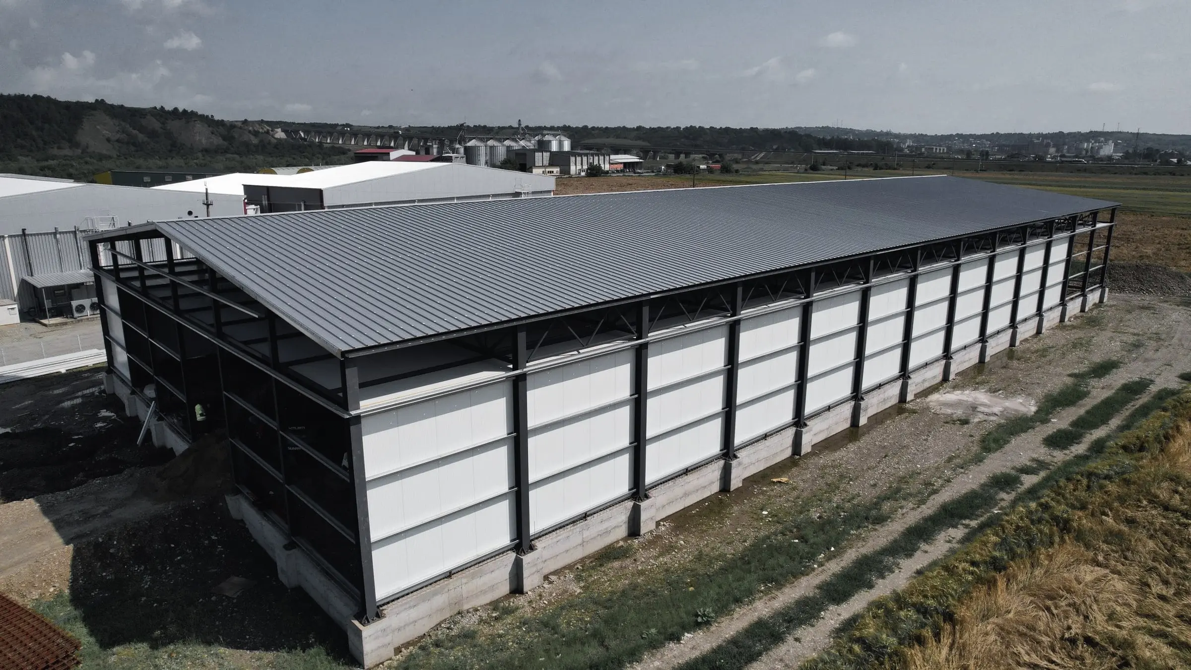 Hală cu structură metalică, acoperiș gri și panouri albe, văzută din dronă