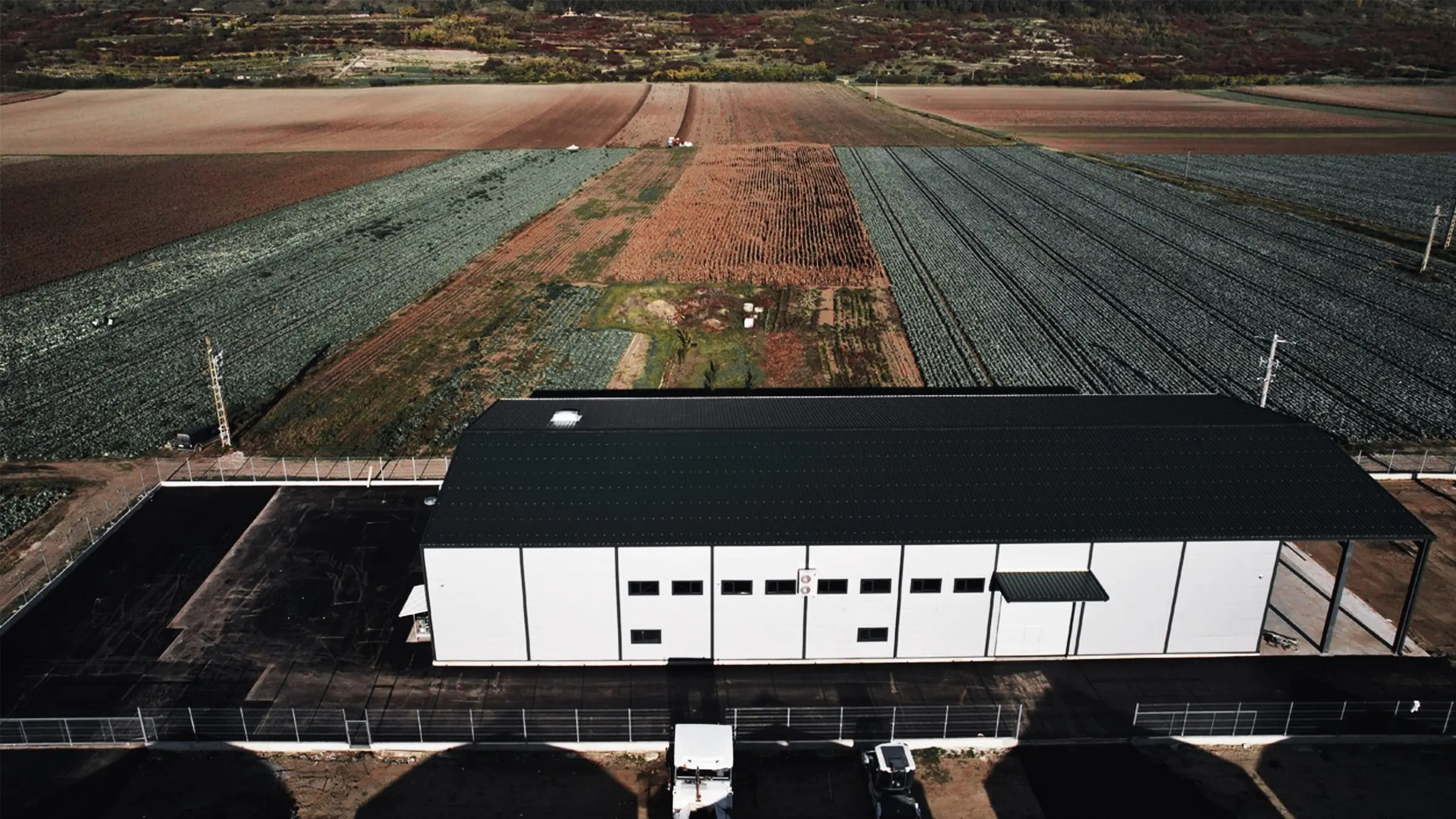 Vedere aeriană a unei hale albe cu acoperiș verde, înconjurată de câmpuri agricole