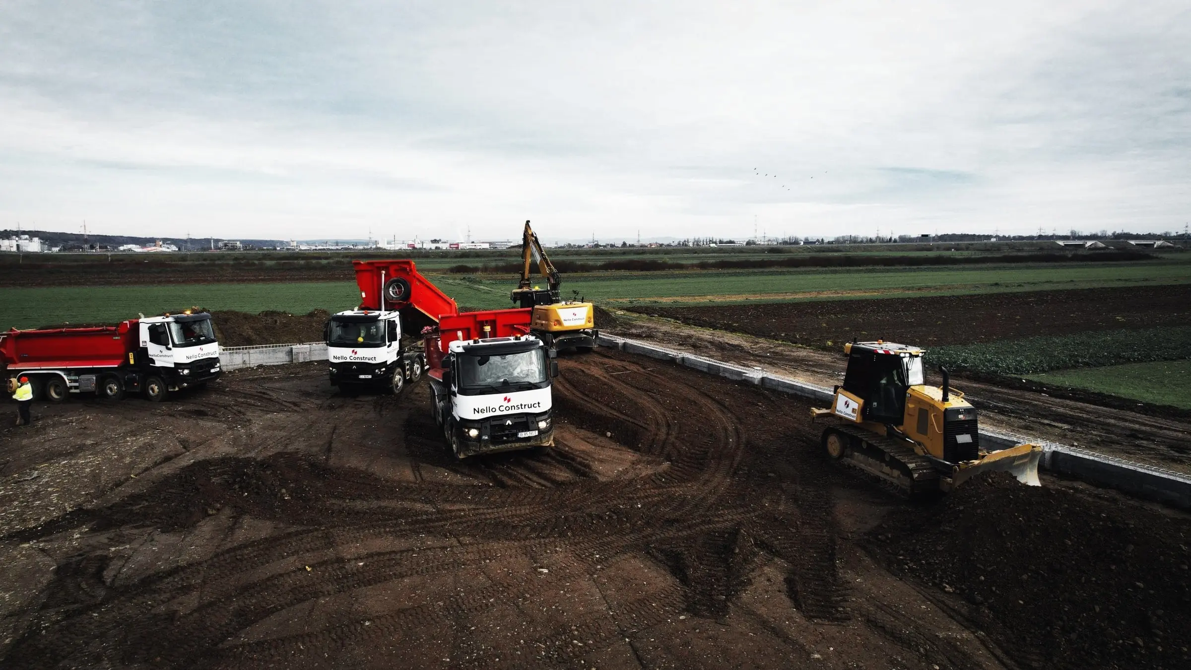 Autobasculante și buldozer lucrează pe un teren decopertat, cu câmpuri agricole în fundal