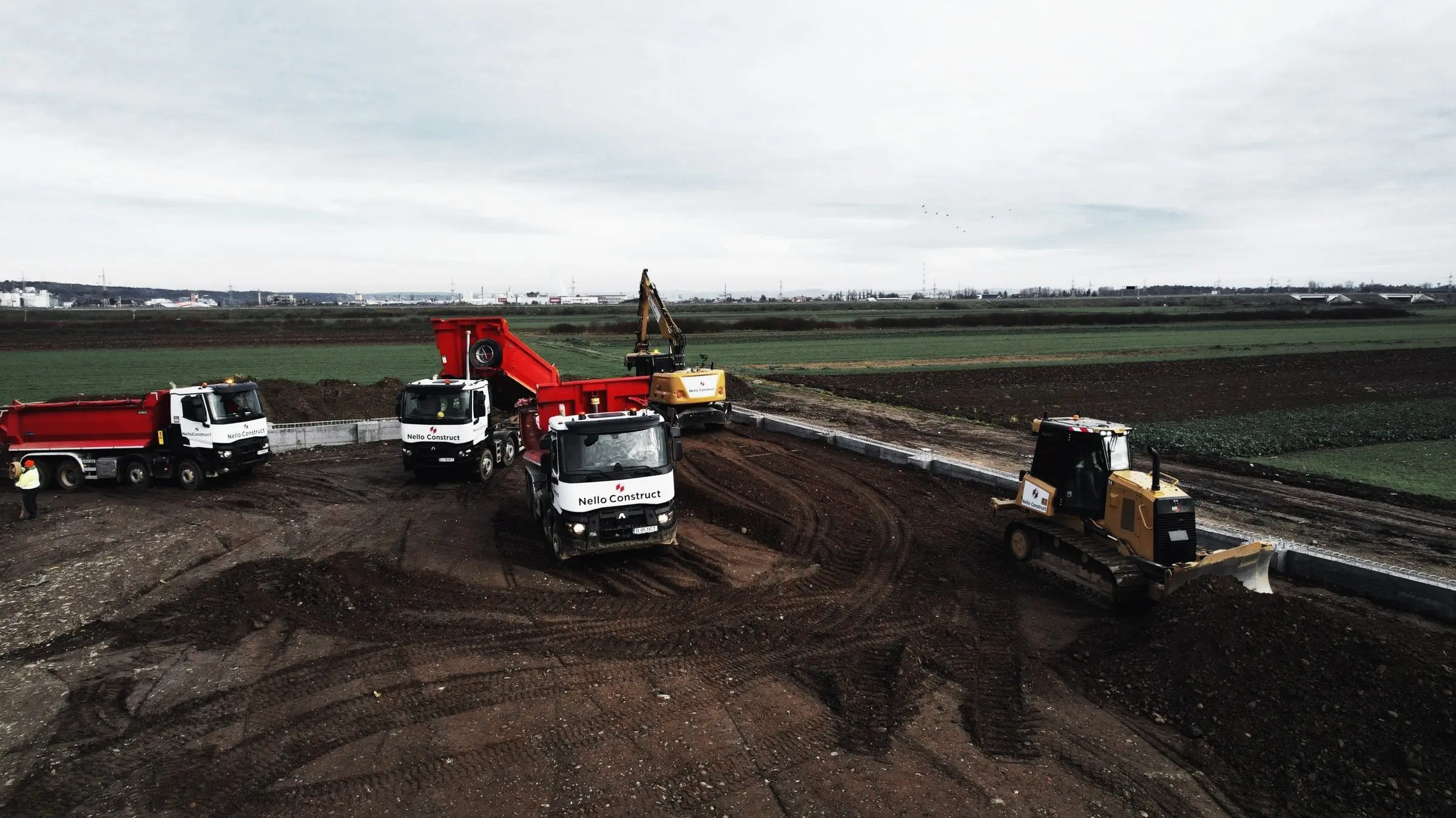 Autobasculante și buldozer lucrează pe un șantier de terasamente, lângă un teren agricol