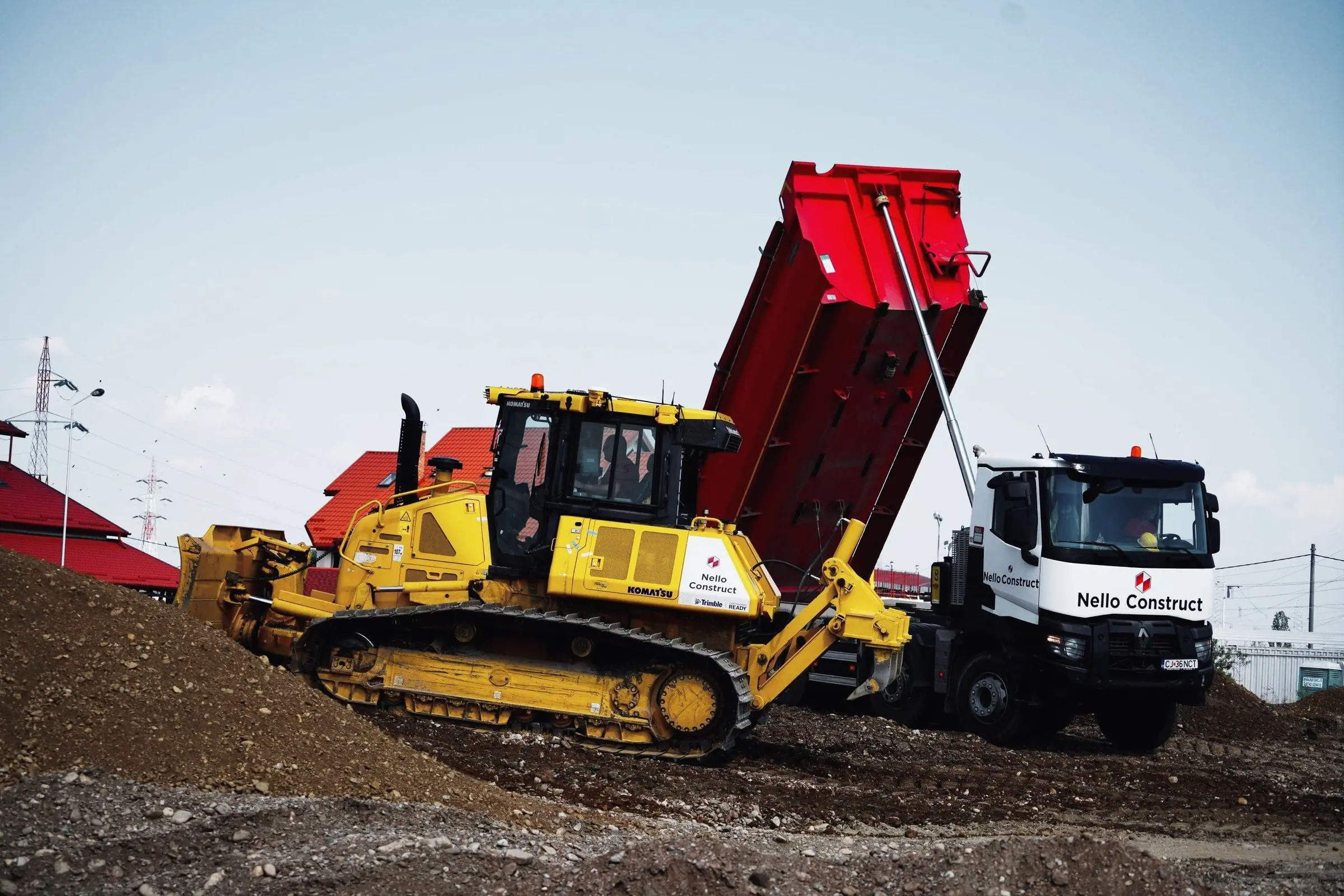 Buldozer galben împinge pământ lângă o autobasculantă cu bena ridicată