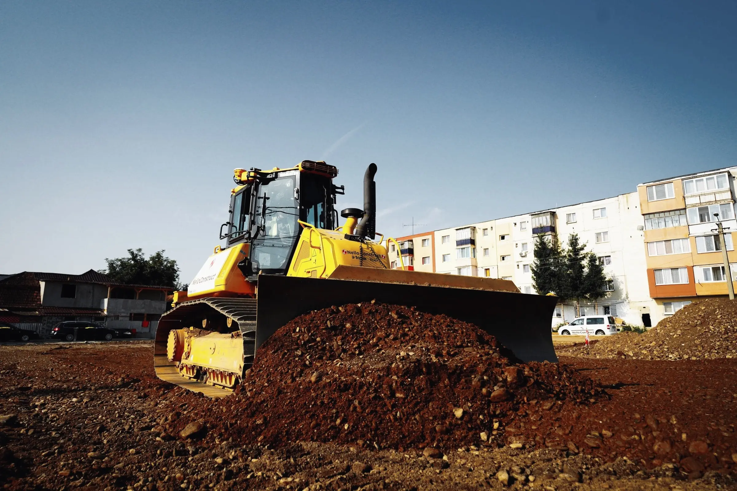 Buldozer galben împinge un morman de pământ, cu blocuri de locuințe în fundal
