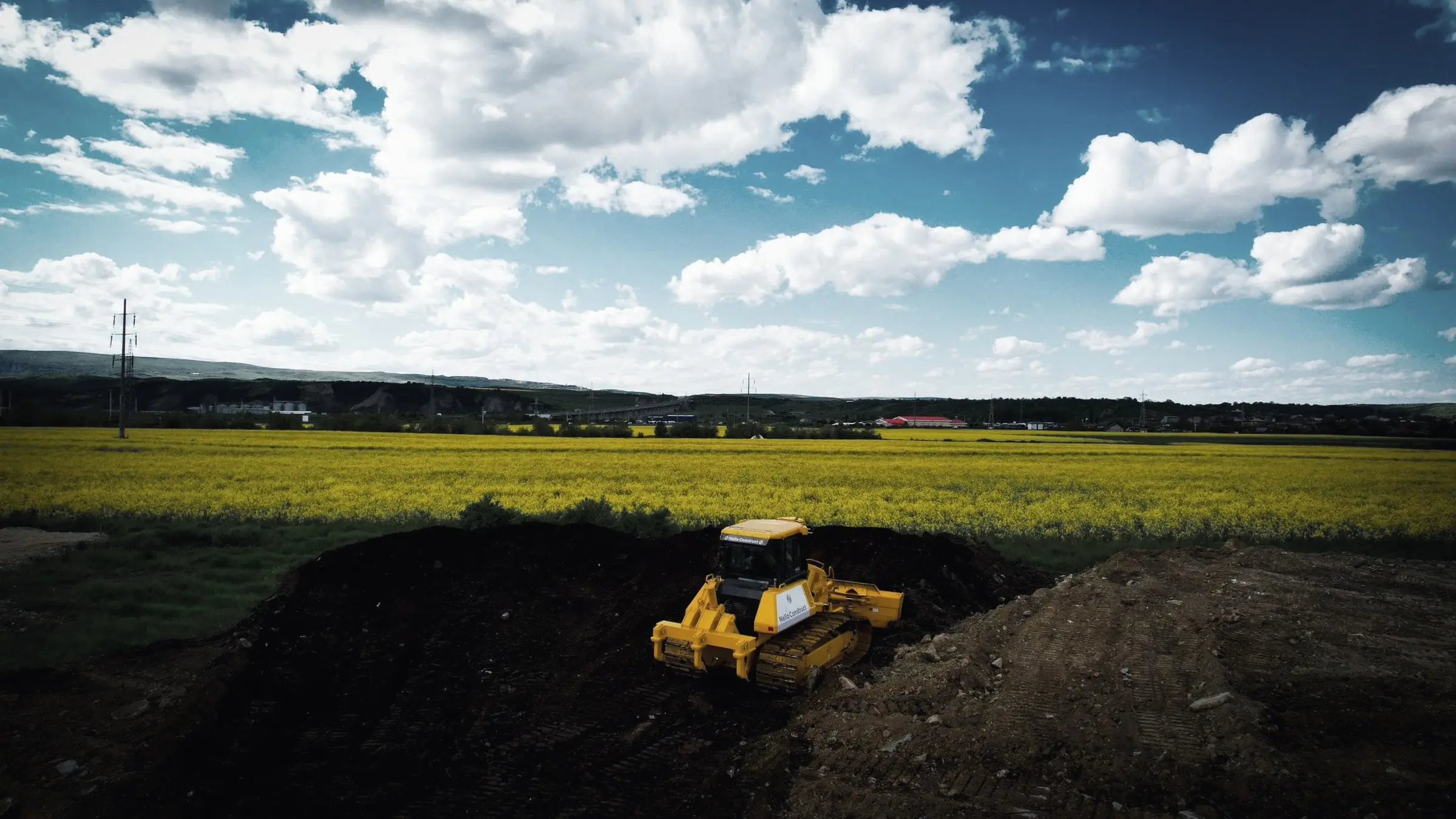Buldozer galben pe un morman de pământ negru, lângă un lan de rapiță