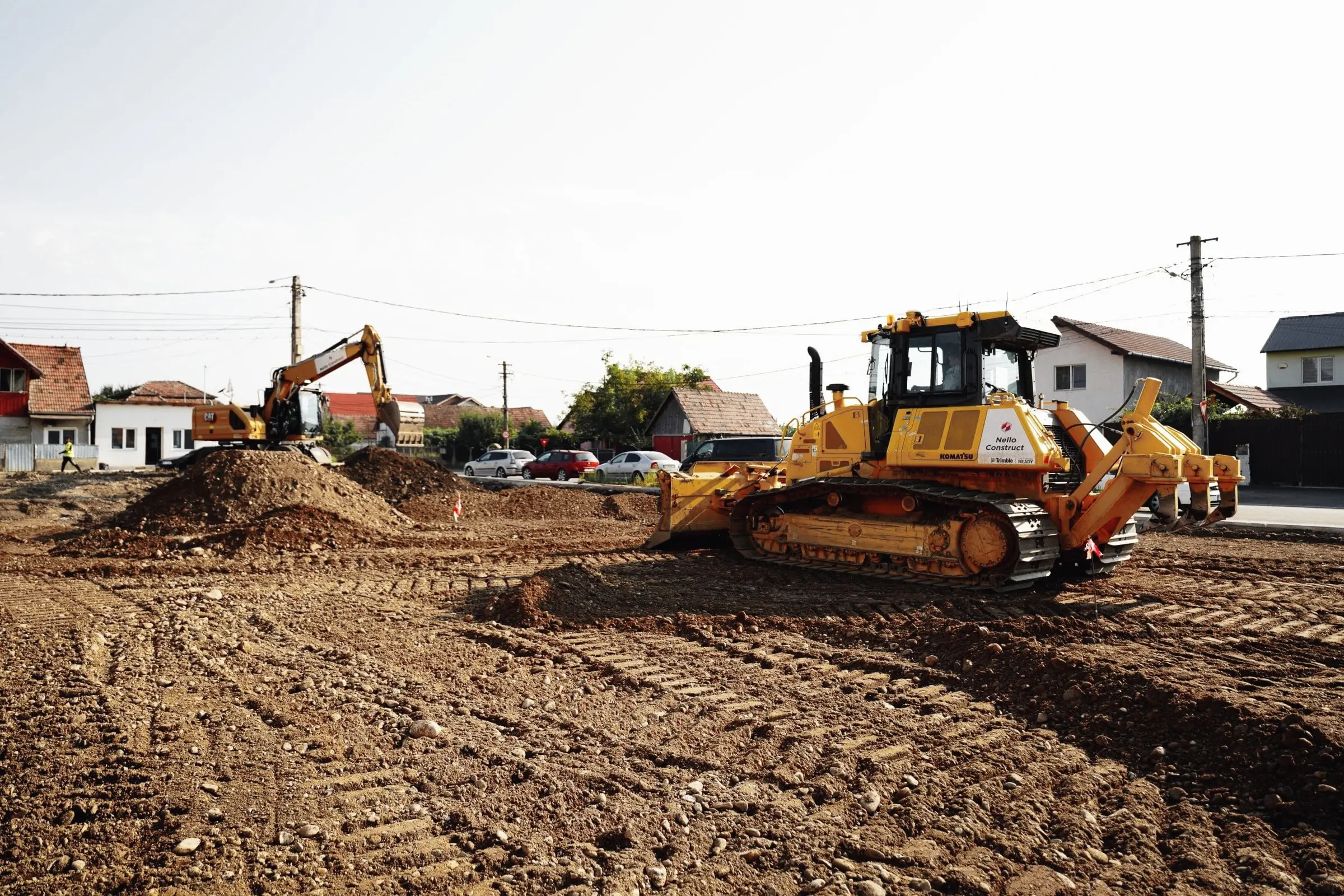Buldozer galben pe teren de pământ, cu case și un excavator în fundal