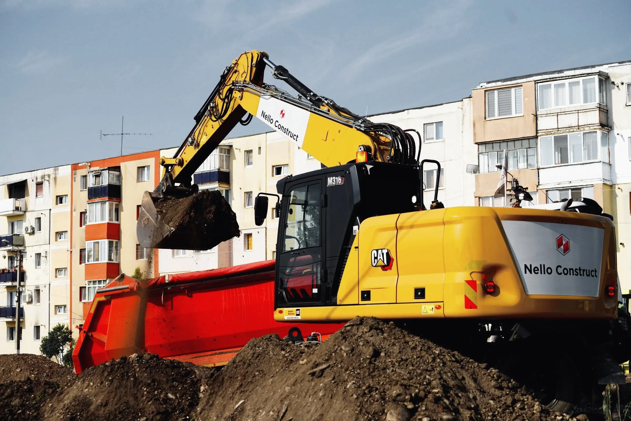 Excavator pe roți încarcă pământ într-o autobasculantă roșie, lângă blocuri