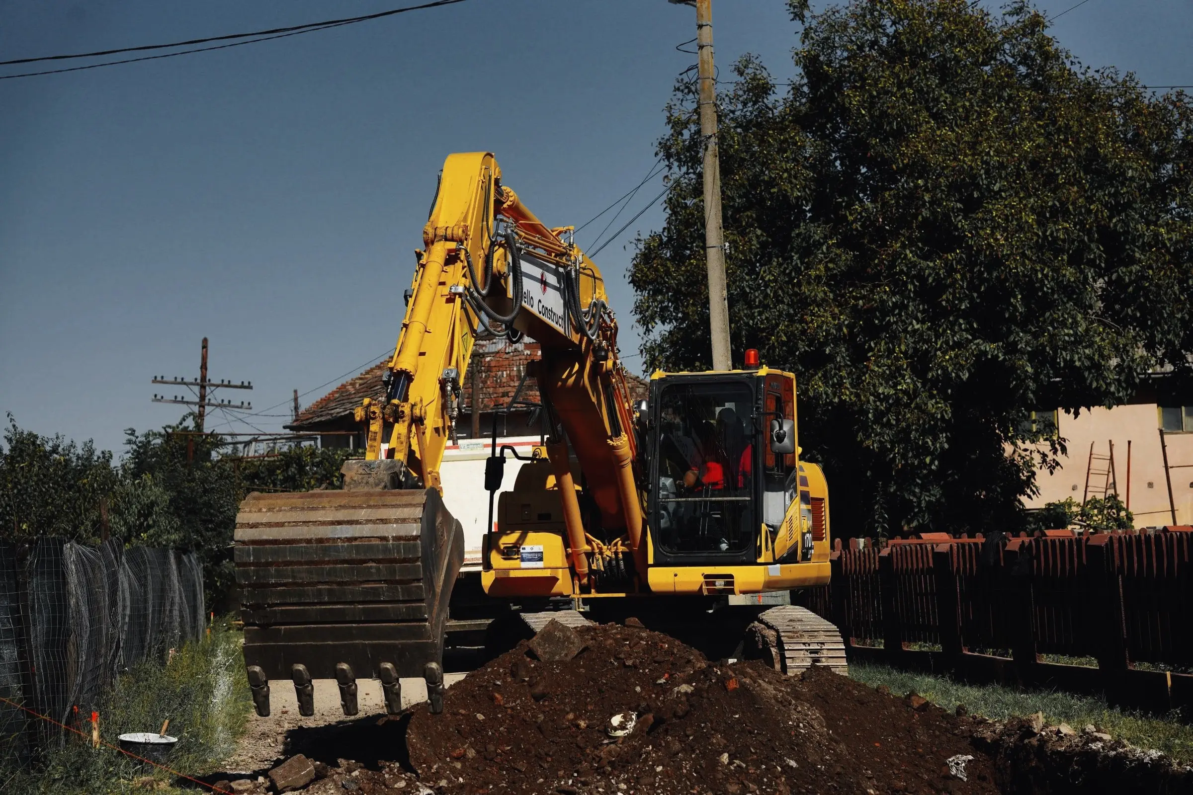 Excavator galben sapă pământ lângă un gard, într-o zonă cu case