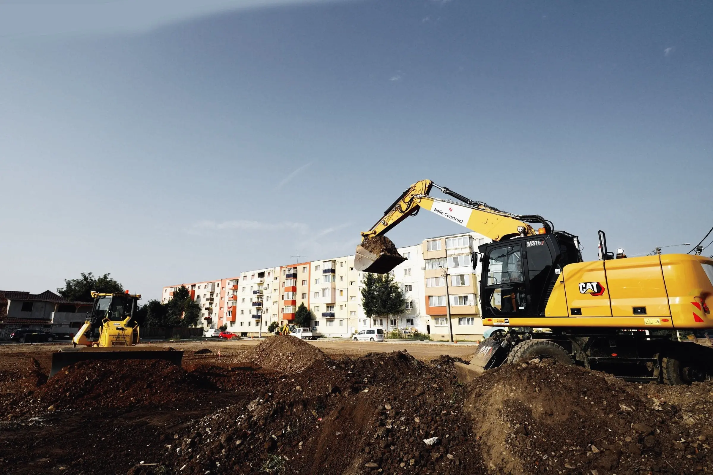 Excavator pe roți lucrează pe un teren de pământ, lângă blocuri de locuințe