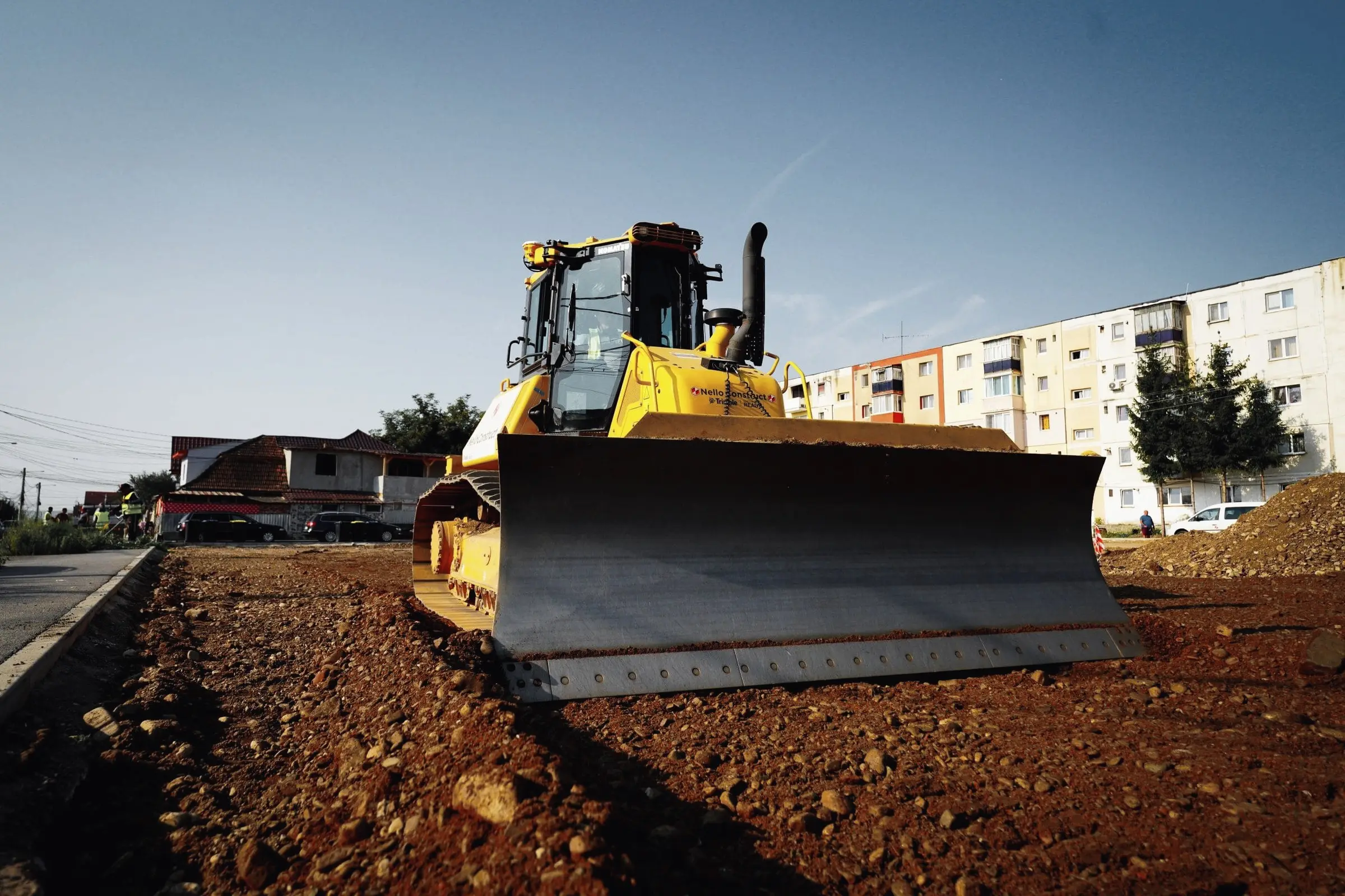 Lama unui buldozer galben pe un teren de pământ, cu blocuri în fundal
