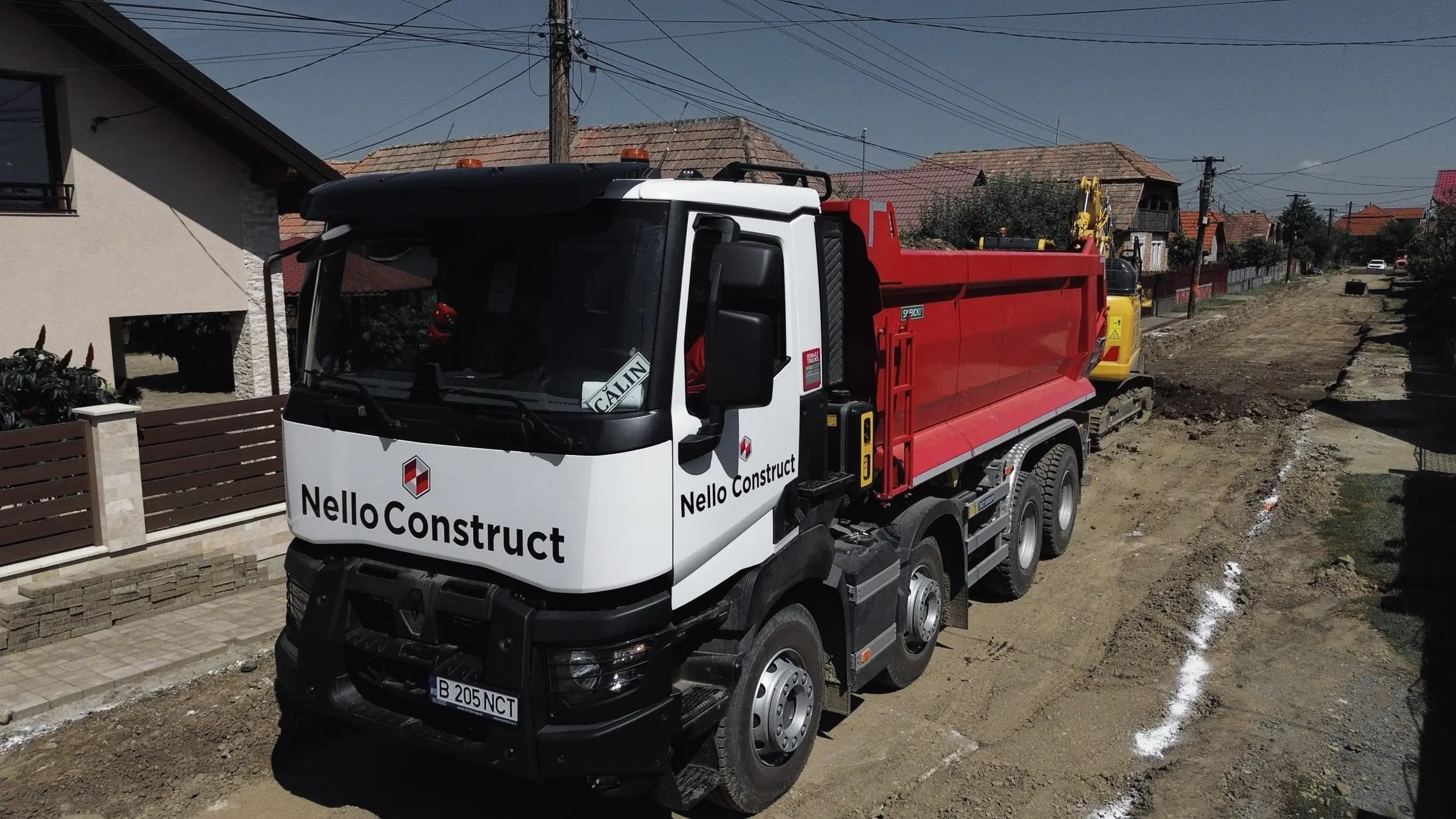 Autobasculantă Nello Construct cu bena roșie pe o stradă de sat în lucru