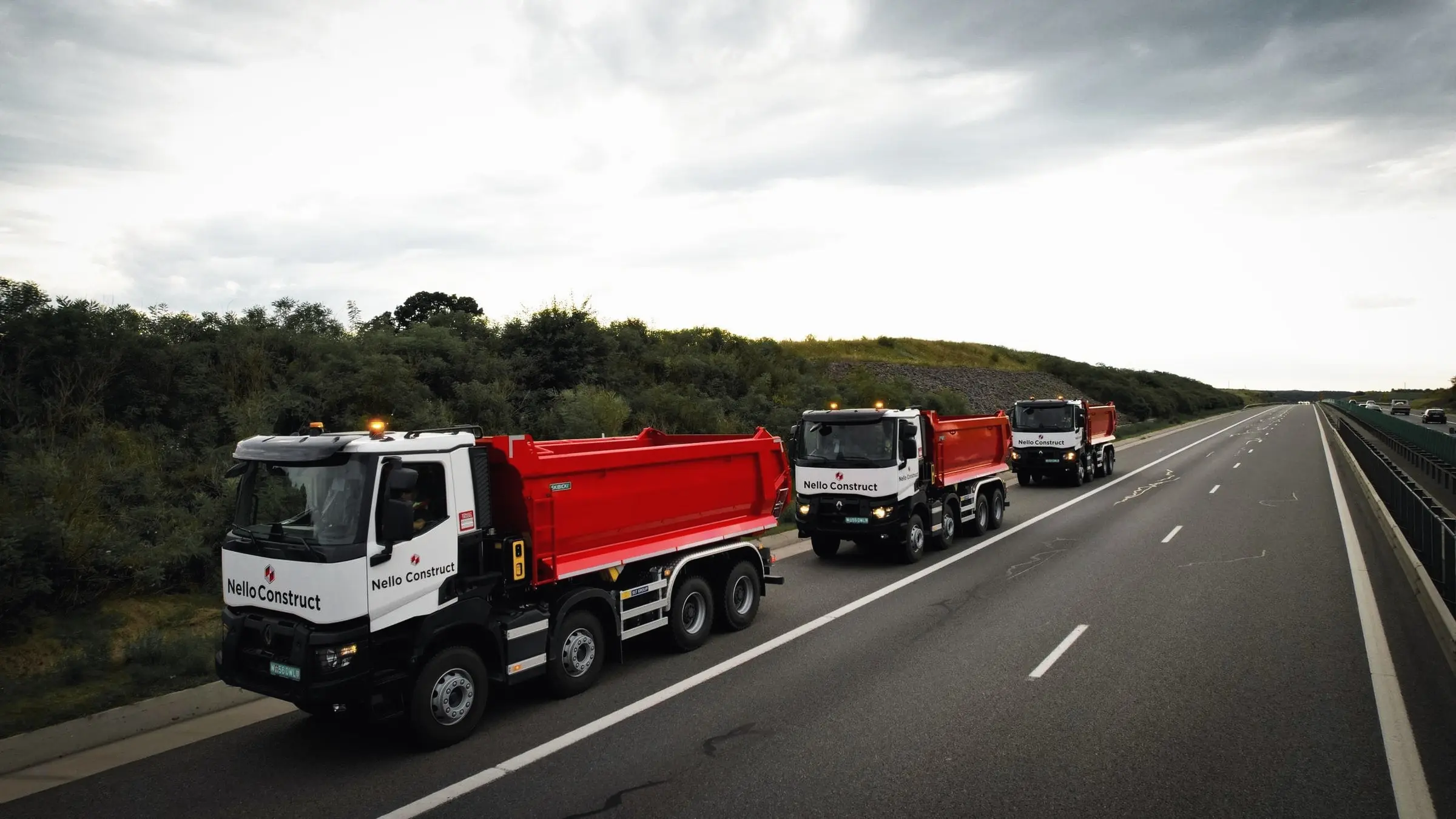 Autobasculante Nello Construct cu bena roșie circulă pe o autostradă, sub cer cu nori
