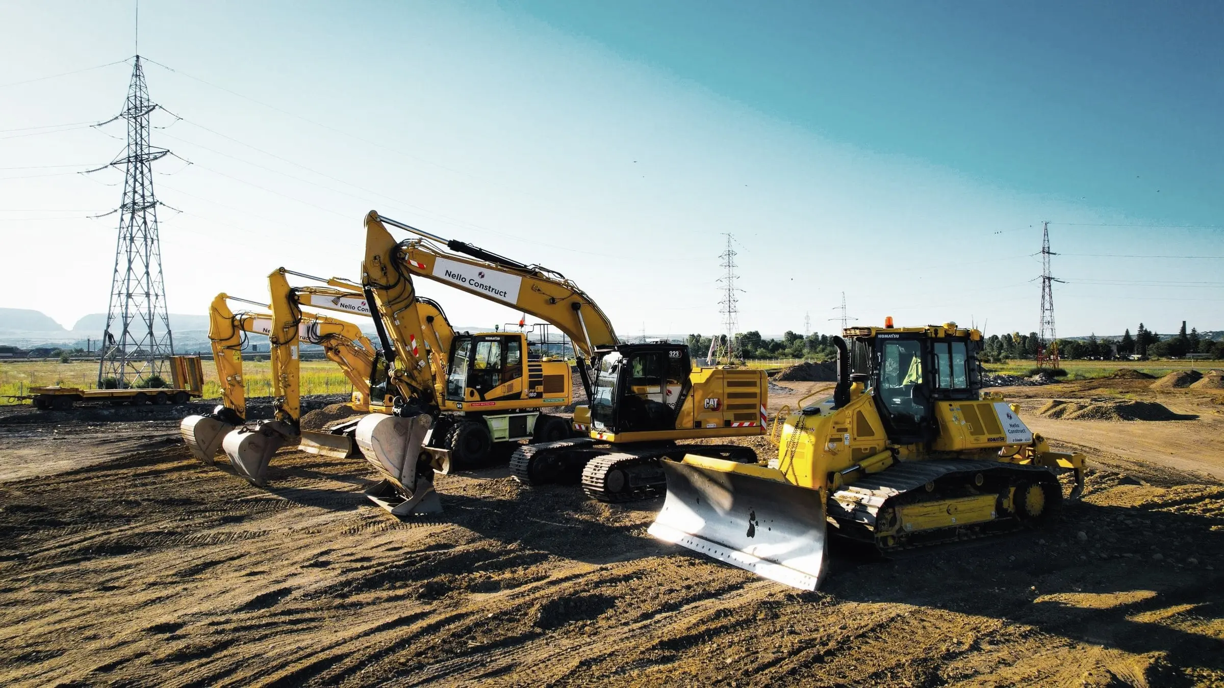 Buldozer și excavatoare galbene aliniate pe teren, cu stâlp de înaltă tensiune în fundal