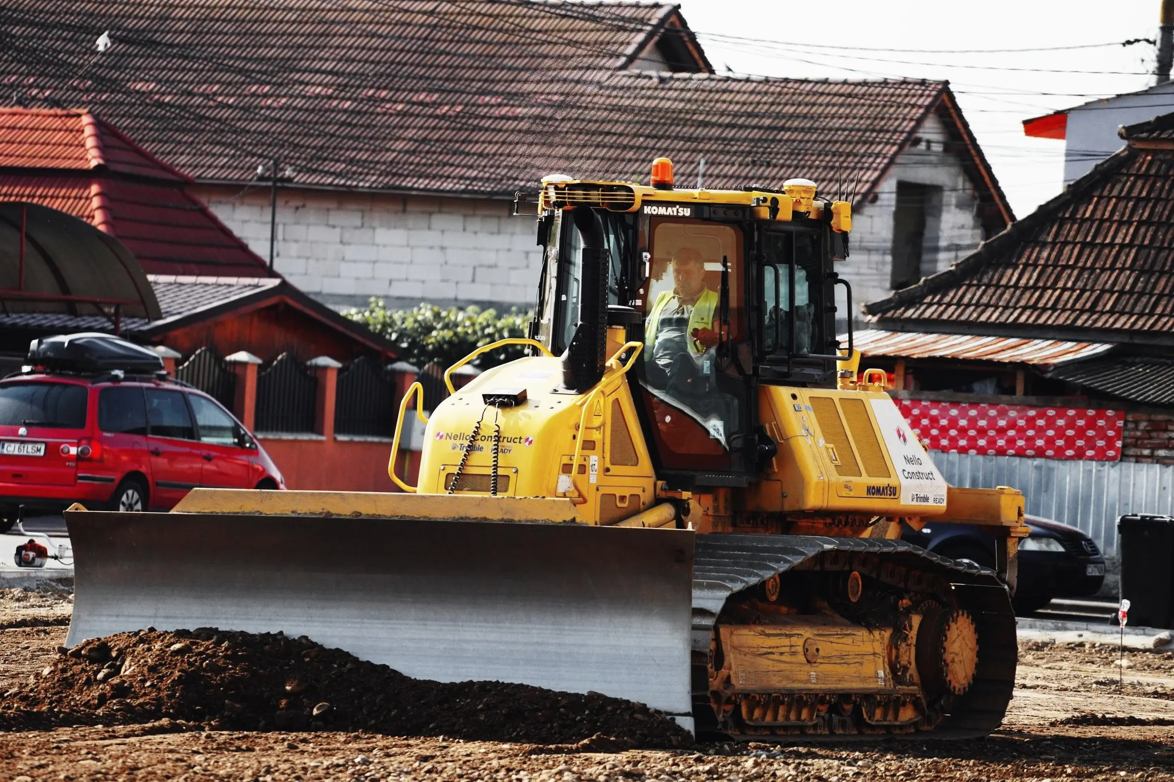 Buldozer galben cu lama jos pe un teren de pământ, în fața unor case