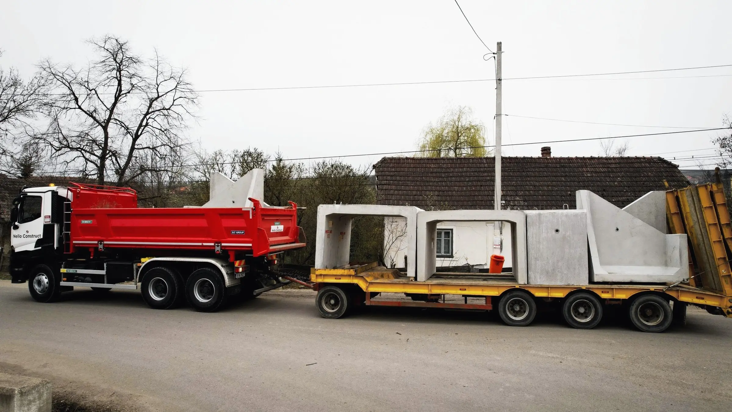 Camion cu remorcă joasă transportă elemente prefabricate din beton pe o stradă de sat