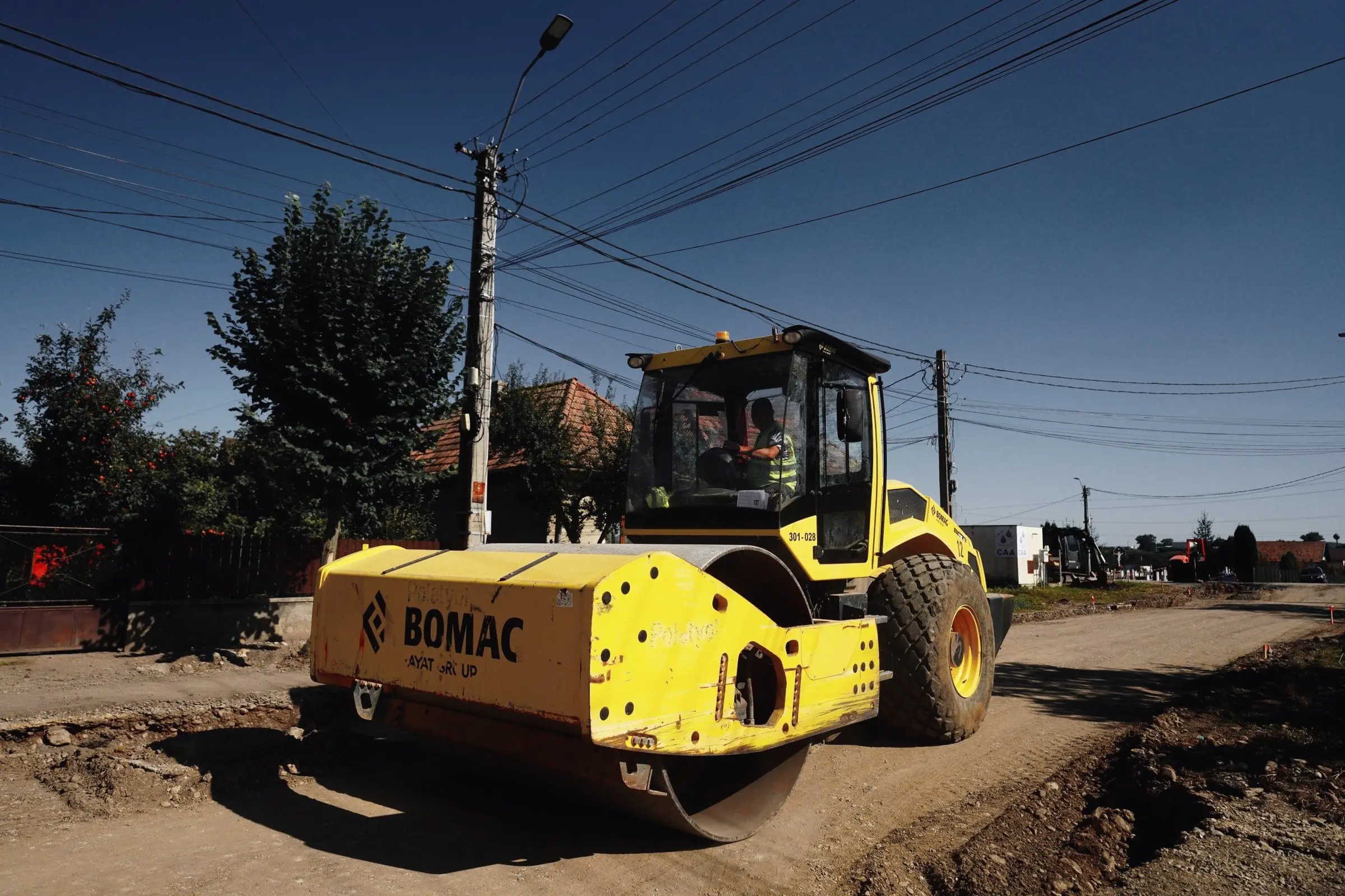 Compactor cu tambur galben Bomac pe un drum de pământ în lucru, lângă case